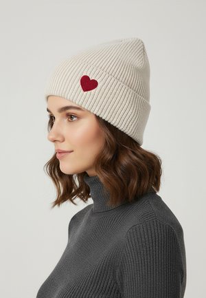 Knit beanie, light beige with ribbed texture, features a small red heart patch on the side. Worn on a model with a gray turtleneck.