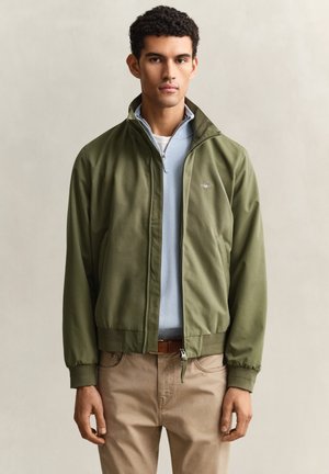 Jonge man draagt een olijfgroene jas met rits over een lichtblauwe trui met halve rits en beige broek, staand tegen een effen achtergrond.