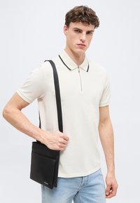 Νεαρός άνδρας που φοράει μπεζ πόλο και ανοιχτόχρωμο τζιν, κρατώντας μαύρη τσάντα χιαστί με λουρί στον ώμο, ουδέτερο φόντο.