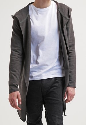 Persoon draagt een donkergrijze open voorkant capuchonvest over een wit t-shirt en zwarte jeans, staand tegen een effen lichte achtergrond.