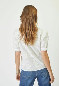Blusa bianca ricamata con motivi a foglia, maniche corte a sbuffo e polsini rifiniti in pizzo. Indossata con jeans in denim blu, che mostrano una vestibilità rilassata.
