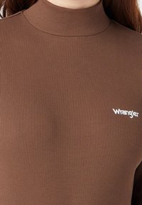Traje de cuello alto acanalado marrón con mangas cortas, con un pequeño logotipo en blanco de "Wrangler" en el área del pecho izquierdo. Textura suave.