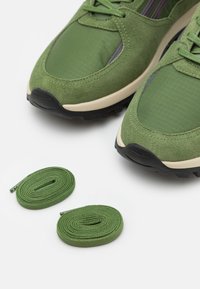 Grüne Wildleder- und Stoffturnschuhe mit schwarzen Sohlen, beigen Zwischensohlen und zwei passenden, spiralförmigen grünen Schuhbändern auf einer weißen Oberfläche.