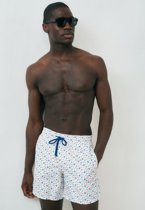 Homme torse nu portant des lunettes de soleil foncées et un short de bain blanc avec un motif d'étoiles multicolores et un cordon bleu, debout contre un mur uni.