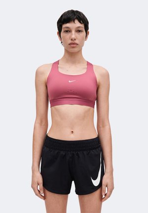 Kobieta nosząca różowy sportowy biustonosz Nike i czarne szorty Nike stoi na jasnym, jednolitym tle.