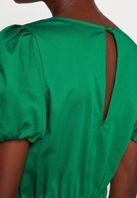 Grön satinklänning med puffärmar, en nyckelhålrygg och elastisk midja, som visar en slät textur och livfull färg.