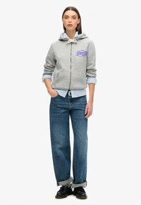 Felpa grigia con zip e tasche frontali, dettagli in testo viola. Indossata sopra una camicia a bottoni blu chiaro e abbinata a jeans larghi blu e scarpe nere.