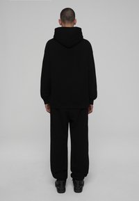 Muž stojící zády oblečený v úplně černém oblečení, včetně mikiny s kapucí, volných kalhot a černých tenisek, na jednobarevném šedém pozadí.