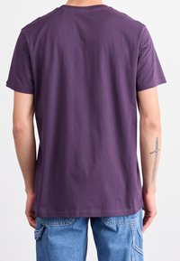 Fialové tričko s krátkým rukávem a kulatým výstřihem, vyrobené z měkkého materiálu. Viditelné švy na lemu a uvolněný střih. Nosené s modrými pracovními kalhotami.