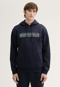 Granatowy kaptur wykonany z miękkiej mieszanki bawełny, z kieszenią kangurką, kapturem ze sznurkiem oraz białym nadrukowanym logo na klatce piersiowej.