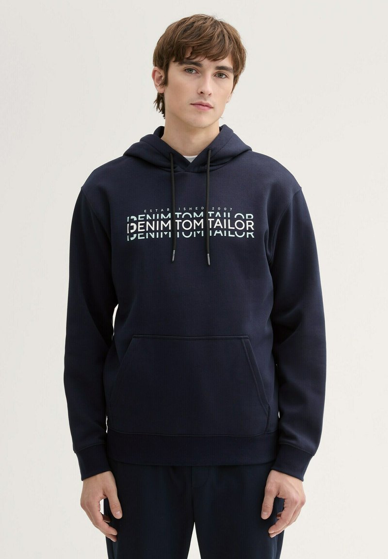 Granatowy kaptur wykonany z miękkiej mieszanki bawełny, z kieszenią kangurką, kapturem ze sznurkiem oraz białym nadrukowanym logo na klatce piersiowej.