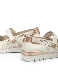 Scarpe bianche in pelle con un design floreale intagliato, accenti metallici dorati, cinturino regolabile e una spessa suola in gomma beige.