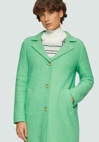 Manteau en mélange de laine vert avec un col à revers, deux poches avant, fermeture à trois boutons et une finition texturée. Col roulé rayé en dessous.
