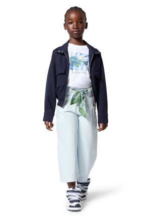 Niña joven con chaqueta azul marino, camiseta blanca con gráfico, pantalones de pierna ancha azul claro con cinturón de bufanda verde y zapatillas azul marino y blancas, de pie.