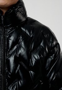 Svart puffjacka med glänsande yta, med hög krage, tjock vaddering och strukturerade detaljer på ytan. Dragkedjestängning.