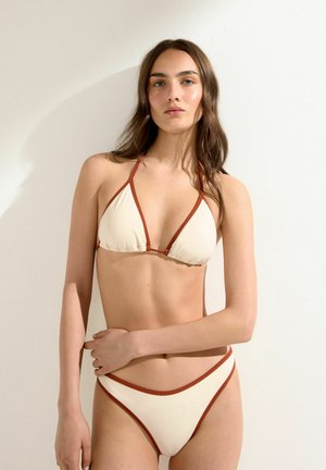 Kvinde iført en cremefarvet bikini med rustfarvede kanter, står op ad en ensfarvet lys væg og ser lige frem med den ene hånd på hoften.