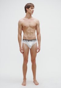 Calzoncillos blancos hechos de tela suave con una cinturilla negra con la marca. El modelo está de pie, descalzo, mostrando un diseño ajustado.