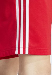 Červené sportovní šortky s hladkou texturou, s třemi bílými pruhy podél boku a olemovaným spodním lemem.