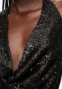 Gros plan sur une robe noire à sequins avec un décolleté en V profond, portée par une personne à la peau foncée avec des mèches de cheveux tressés visibles.