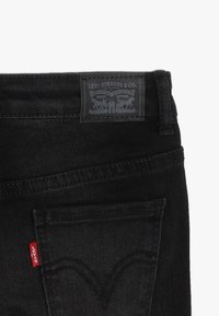 Pantalones vaqueros negros de mezclilla con un parche de cuero con el logo que dice "Levi Strauss & Co." y una pestaña roja en el bolsillo lateral. Presentan costuras curvas en los bolsillos.