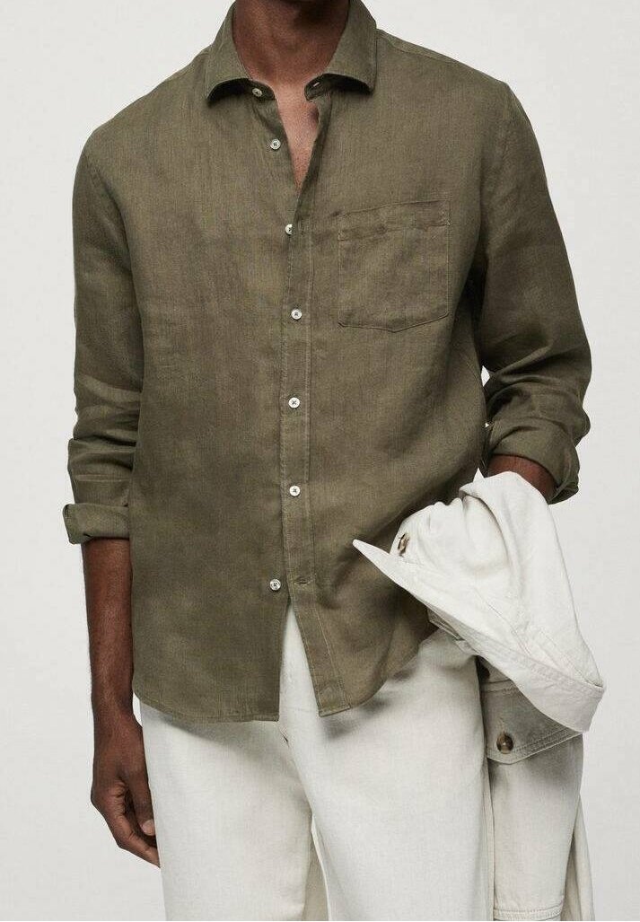 Homme portant une chemise boutonnée en lin vert olive avec une poche poitrine, tenant une veste blanche, associée à un pantalon blanc, sur un fond neutre.