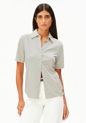 Vrouw met lang donker haar, die een wit kort mouwshirt met dunne zwarte horizontale strepen en een witte broek draagt, tegen een effen achtergrond.