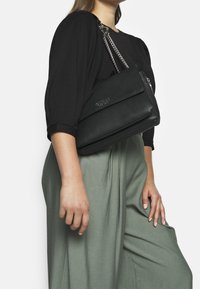 Borsa a mano nera in pelle con tracolla in catena argentata, caratterizzata da una patta frontale e una superficie texturizzata. Il modello indossa un top nero e pantaloni verdi.
