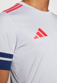 Vit träningsskjorta med röda tre-randiga detaljer på axlarna, marinblå ärmar med band och en röd Adidas-logotyp på framsidan. Slät textur.