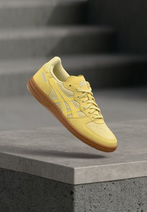 Zapatilla amarilla con suela marrón flotando sobre un borde de concreto, con escaleras desenfocadas al fondo.
