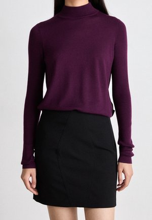 Femme portant un pull à col roulé violet moulant à manches longues et une jupe noire taille haute sur un fond uni.