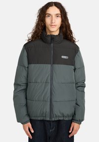 Giovane con lunghi capelli ricci che indossa un giubbotto invernale nero e grigio scuro con zip frontale e logo sul petto.