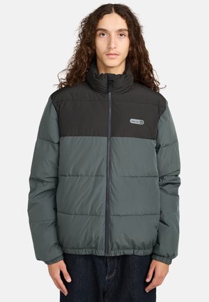 Giovane con lunghi capelli ricci che indossa un giubbotto invernale nero e grigio scuro con zip frontale e logo sul petto.