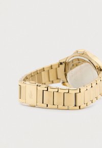 Gold metal link watch band with clasp featuring a crocodile logo, attached to a round watch case with visible back cover.