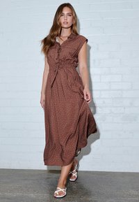 Brown polka dot midi dress with a button-down front, short sleeves, elastic waist, and flowing fabric. Worn with white sandals.