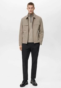 Beige jas met twee voorzakken, gelaagd over een lichtgekleurde overhemd met hoge kraag, gecombineerd met zwarte broek en zwarte schoenen.