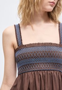 Γυναίκα που φορά καφέ φόρεμα χωρίς μανίκια με μπλε κεντημένα μοτίβα διαμαντιού και πλέγματος στο μπούστο, κομμένο στο ύψος του λαιμού και των ώμων.