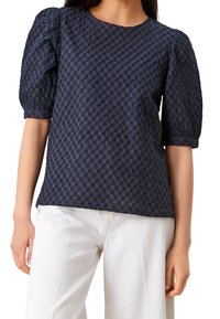 Top puff-sleeve blu navy in tessuto testurizzato con piccoli motivi ricamati, scollatura rotonda e vestibilità comoda. Abbinato a pantaloni chiari.