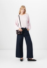 Woman wearing a light pink sweater, wide-leg dark blue jeans, black flats, and a small dark brown crossbody bag, standing with one hand in pocket.