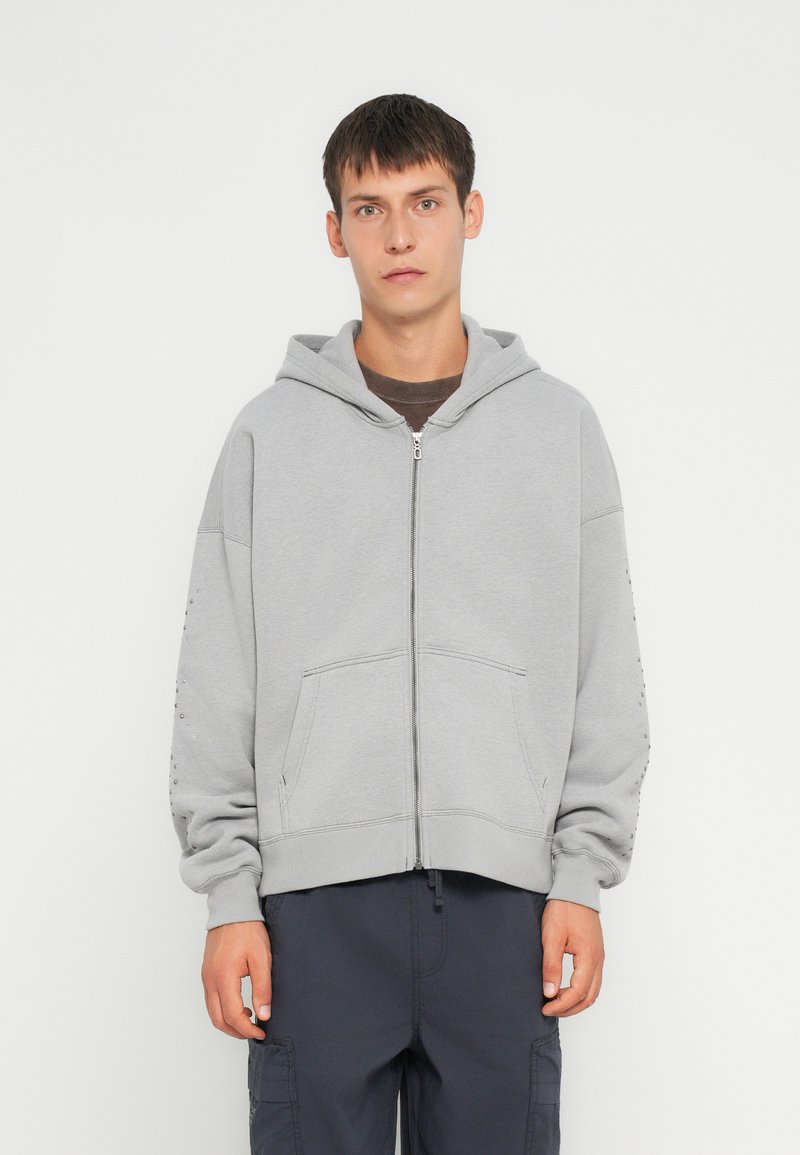 Felpa grigia con zip, cappuccio, tasche a marsupio e polsini a coste. Il tessuto sembra morbido con una texture liscia e un minimo di dettagli sulle maniche.