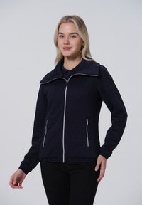 Veste matelassée bleu marine avec col montant, fermeture éclair à l'avant et deux poches latérales. Présente des motifs triangulaires et des poignets élastiques.
