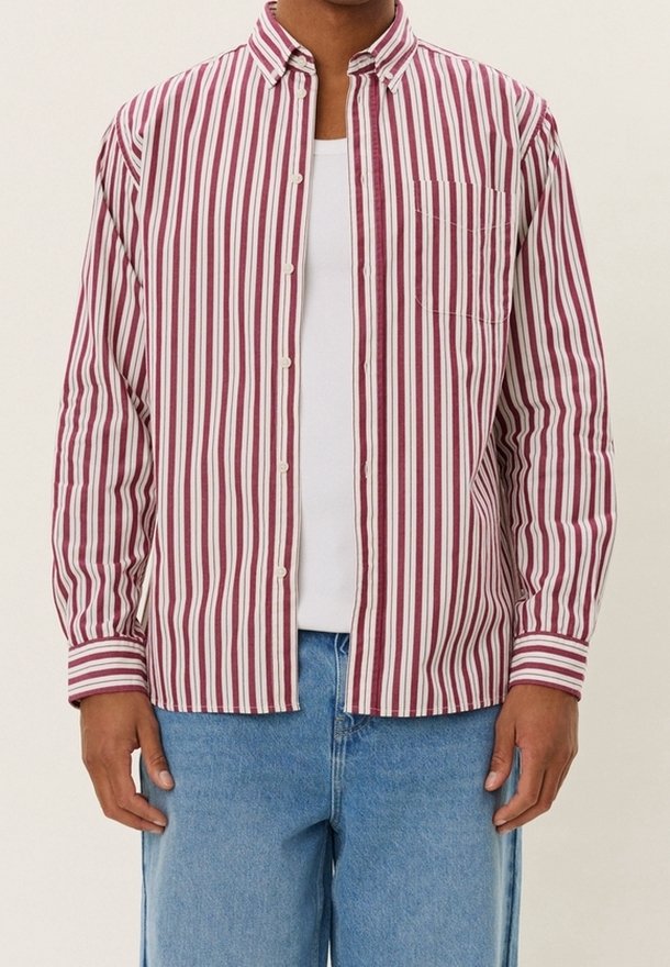 Homme portant une chemise à boutons rayée verticale rouge et blanche par-dessus un t-shirt blanc et un jean bleu clair, debout les mains détendues.
