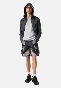 Graue Zip-Up-Jacke mit Kapuze, hellgrauer Pullover und schwarze Shorts mit geometrischem Muster. Schwarze Sportschuhe und weiße Socken runden das Outfit ab.