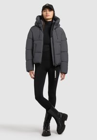 Pelēks polsterēts jaka ar kapuci, ar rāvējslēdzēja aizdari un izteiktu šuvi, saskaņota ar melniem legingiem un masīviem melniem zābakiem.