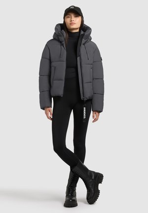 Grijze gewatteerde jas met een capuchon, ritssluiting en opvallende stiksels, gecombineerd met zwarte leggings en stevige zwarte laarzen.