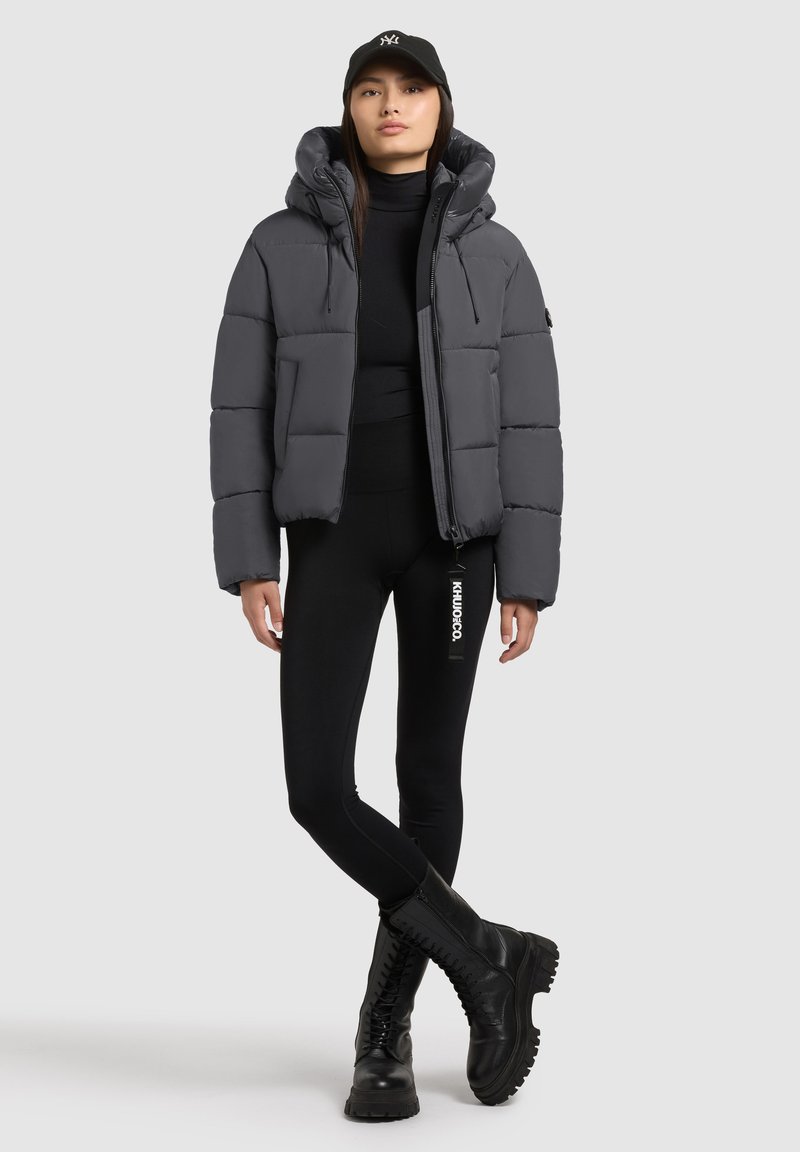 Pelēks polsterēts jaka ar kapuci, ar rāvējslēdzēja aizdari un izteiktu šuvi, saskaņota ar melniem legingiem un masīviem melniem zābakiem.