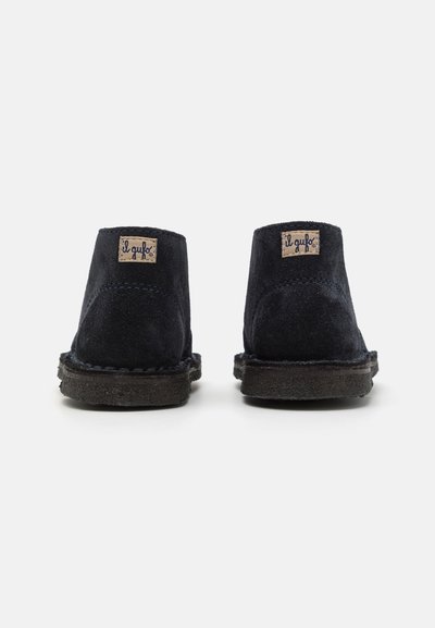 Botine din suede bleumarin cu un aspect moale, având vârful rotund, detalii cusute și un etichetă cu logo pe spate.