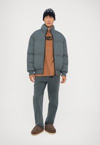 Grå ribbad pufferjacka, brun grafisk t-shirt, grå byxor och mörkbruna tofflor med mönster. Stylad med en stickad svart och grå mössa.