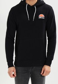 Hombre vistiendo una sudadera negra de ellesse con cordones blancos y bolsillo frontal, combinada con jeans gris oscuro.