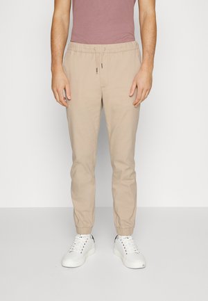 Hombre con pantalones jogger beige con puños elásticos y cintura con cordón, zapatillas blancas y una camiseta de manga corta color malva, de pie contra una pared blanca.