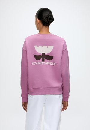 Femme avec les cheveux attachés en chignon portant un sweat-shirt rose avec une fleur stylisée et le texte "Scandinavian community" au dos, associée à un pantalon blanc.